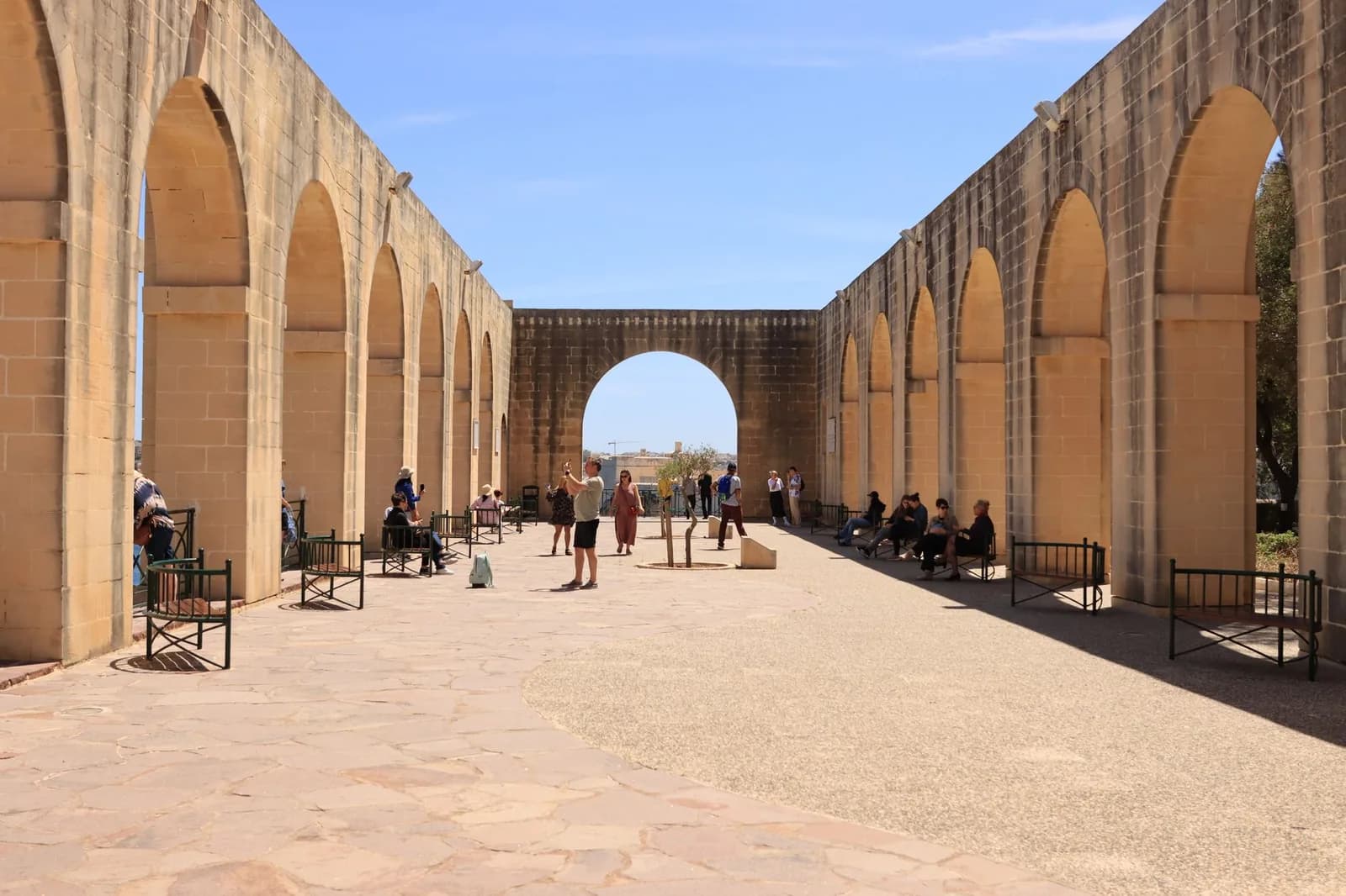Longues arcades en pierre dorée sur les remparts de La Valette, sous un ciel bleu, avec des visiteurs sur l'esplanade