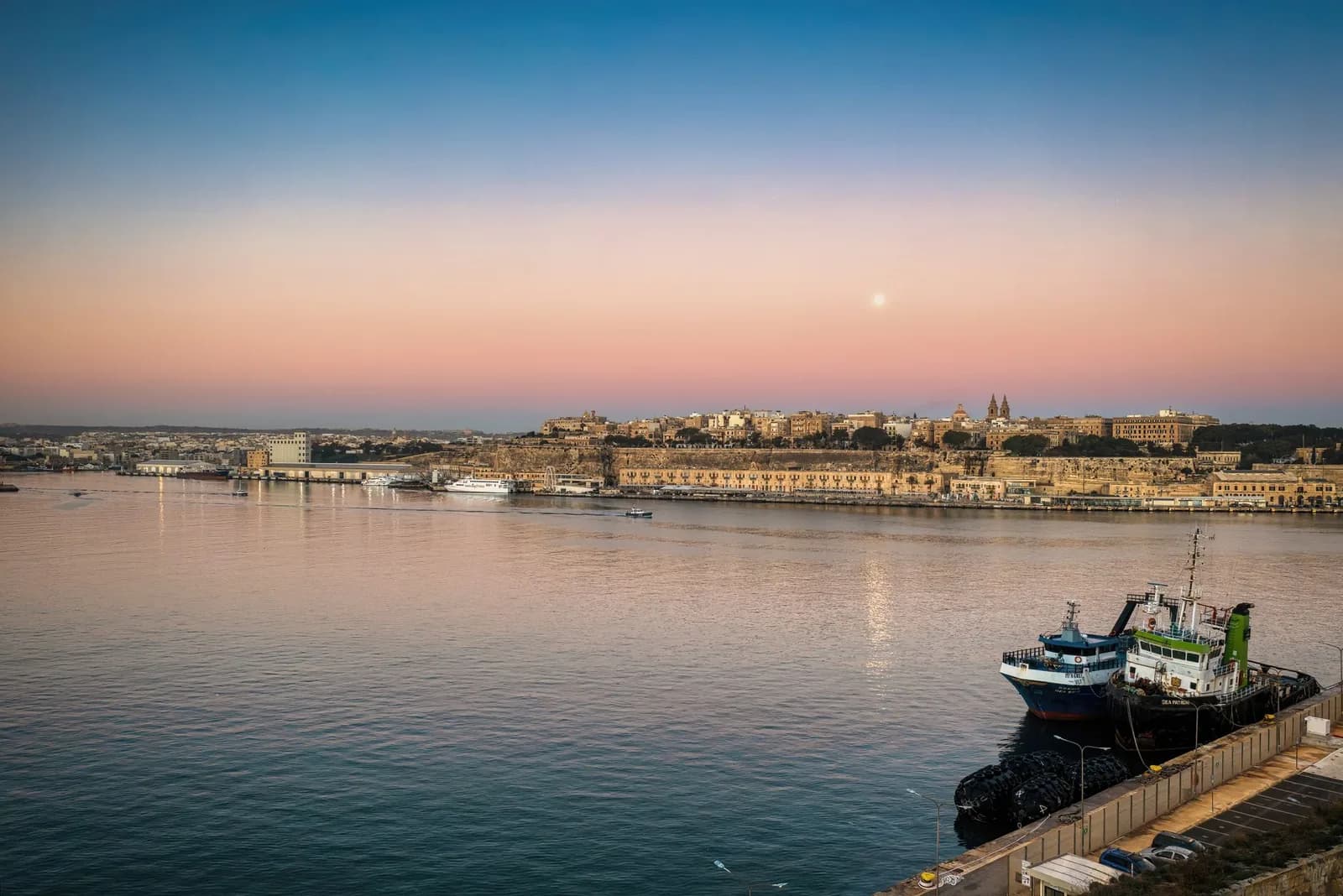 Rade de Malte au crépuscule, ciel rose au-dessus des remparts et de bateaux de pêche amarrés