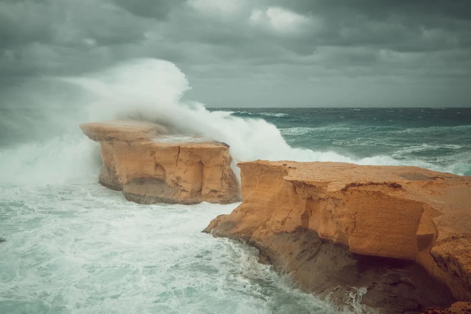 Grosse vague s'écrasant sur des plateaux rocheux de calcaire ocre, sous un ciel gris et couvert