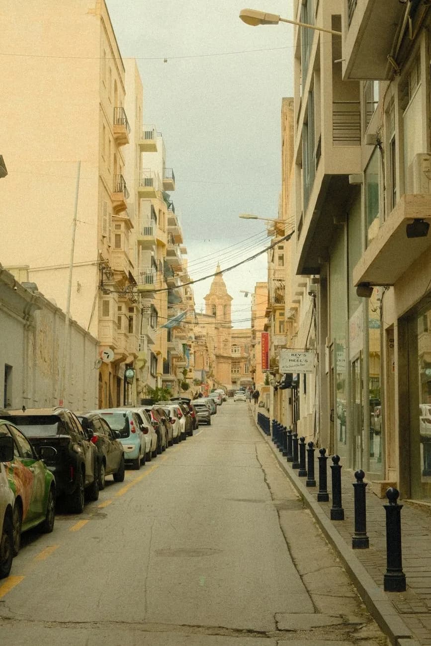 Rue commerçante de Sliema à Malte bordée de boutiques et de voitures garées, avec un clocher en arrière-plan