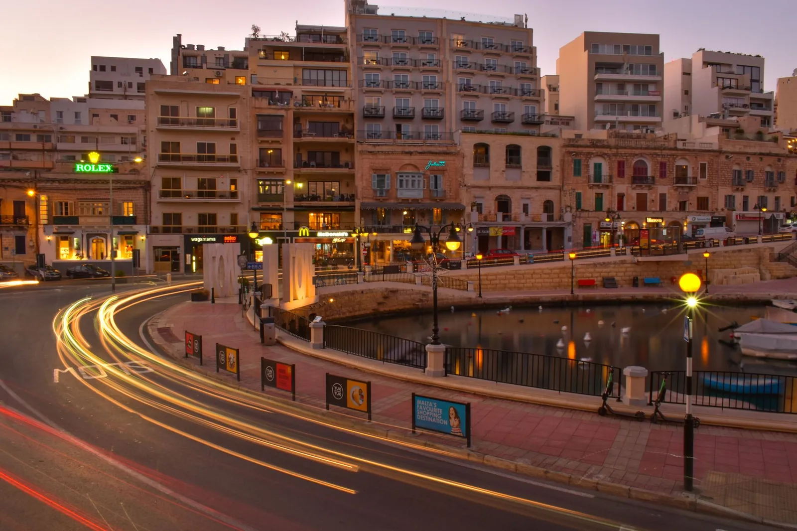 Spinola Bay à St Julian's, Malte, à la tombée de la nuit : bars éclairés le long du port et traînées lumineuses des voitures
